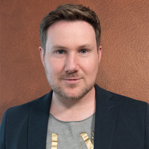 Auf diesem Bild sieht man den Zauberer Axel Dedering. Der Hintergrund ist rostbraun und Axel Dedering trägt ein Jacket. Er lächelt.